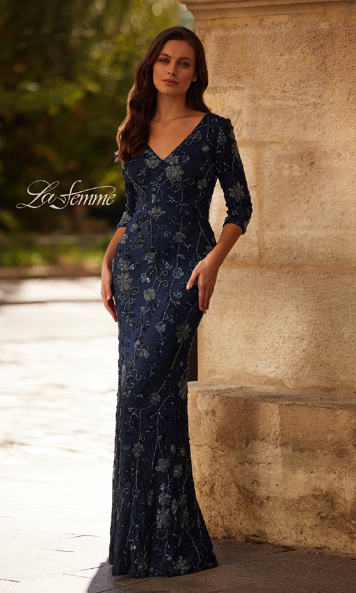 A woman stands outdoors against a stone wall, wearing the Navy Blue Beaded Formal Gown: La Femme 33092, a floor-length fitted dress with floral embroidery and three-quarter sleeves.